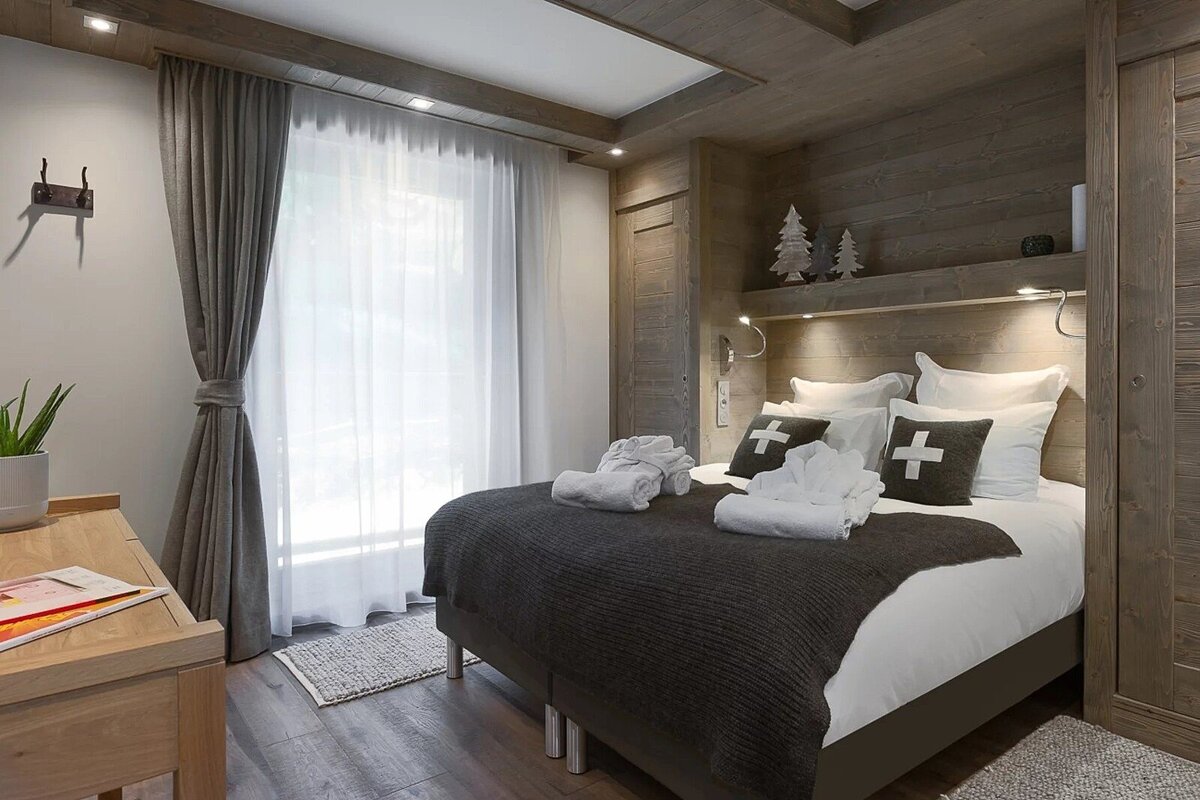 A cozy, modern chalet-style bedroom with wood-paneled walls, a bed featuring cross-patterned pillows, a bright window, and warm lighting.