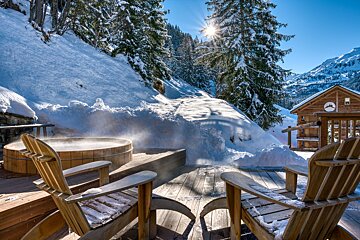 A wooden hot tub steams on a snowy deck with two Adirondack chairs, set against a sunny, snow-covered mountain scene with pine trees and a distant cabin.