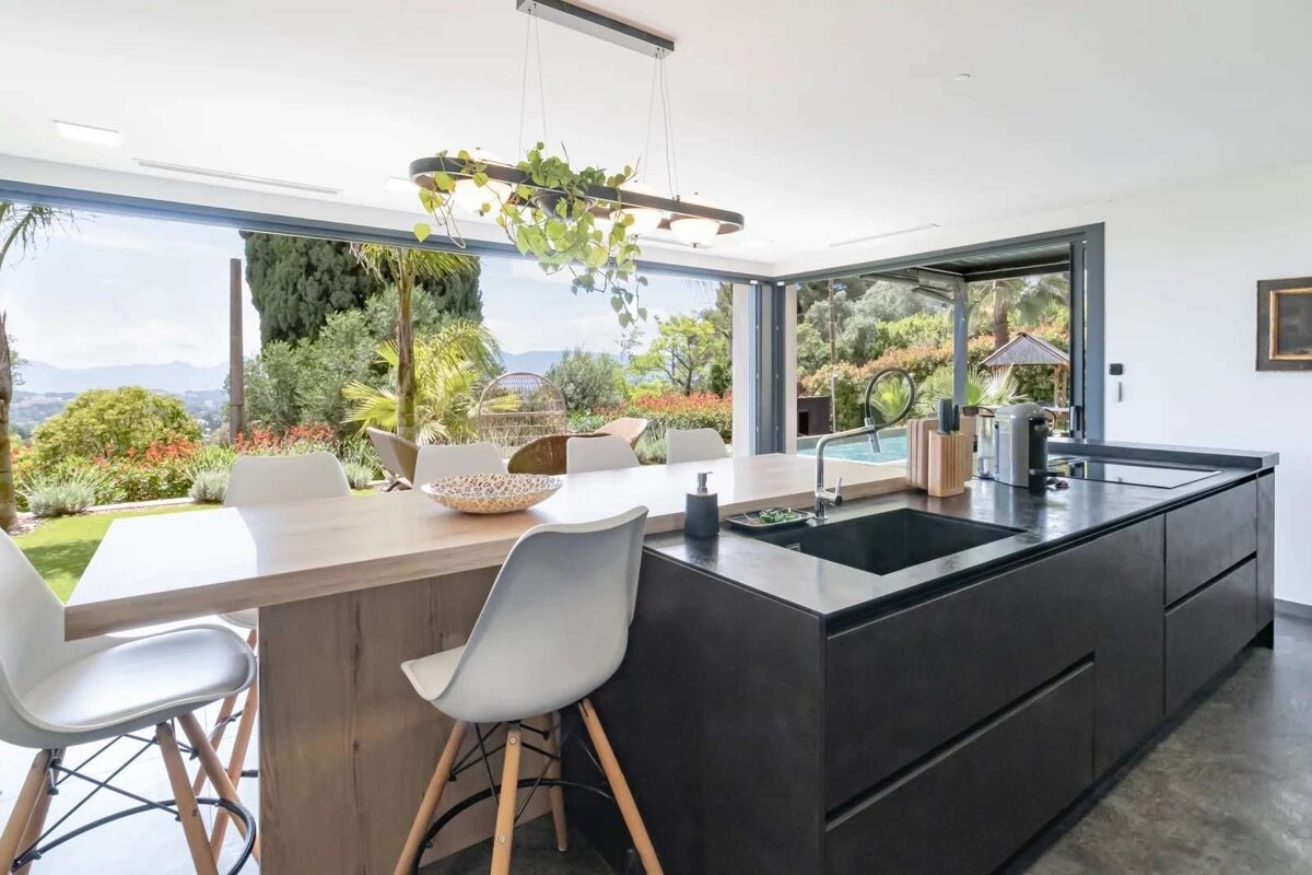 Sleek kitchen and dining space with an integrated table, featuring large glass doors that open to a beautiful garden, pool, and scenic view.