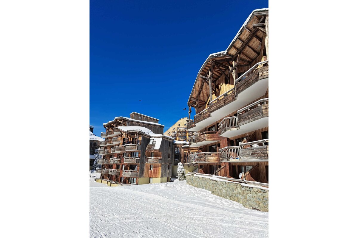 Snow-covered wooden chalets with balconies stand tall against a clear blue sky, surrounded by pristine snow, indicating a winter ski resort.