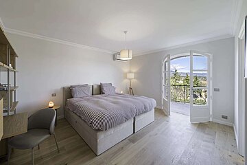 Bright bedroom with a striped bed, wooden floors, and French doors opening to a balcony with a scenic view of hills and trees.