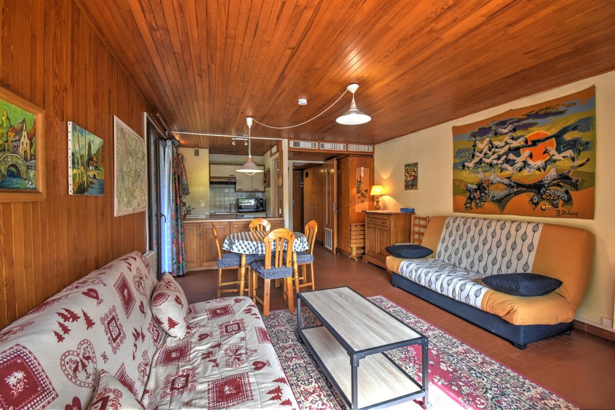 A warm, wood-paneled living/dining room with two sofas, a dining table, and a kitchen view. Features eclectic art and a cozy, rustic ambiance.