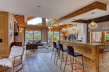 Open-plan modern chalet living room with a wooden bar, stools, sofa, large windows, and contemporary lighting throughout.