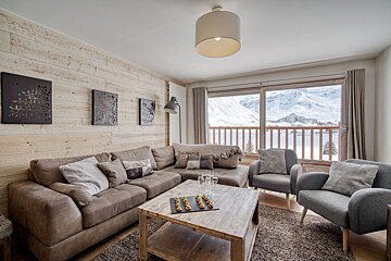 A cozy, modern living room with a large couch, wood-paneled walls, and a panoramic window offering stunning views of snowy mountains and a resort.