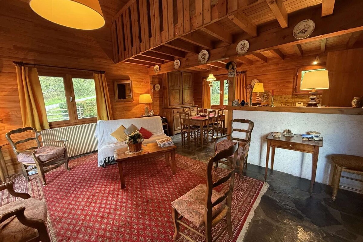 A cozy, rustic log cabin interior combining living and dining areas. Features warm wooden walls, a red rug, sofa, and wooden furniture under soft lighting.