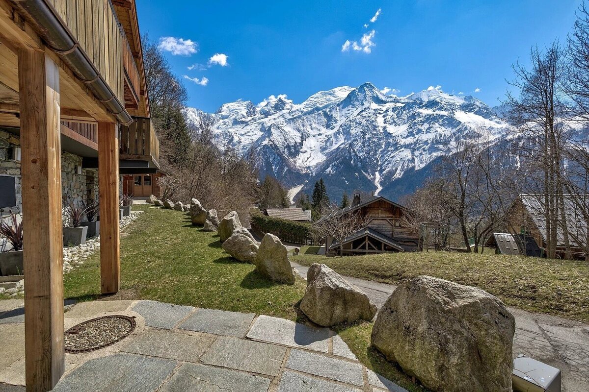 A sunny mountain view featuring snow-capped Mont Blanc, a wooden chalet, and a stone patio leading to a grassy slope with large rocks.