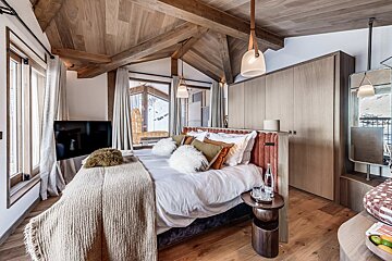 Cozy alpine chalet bedroom with a vaulted wooden ceiling, plush bedding, and snowy mountain views through the windows.