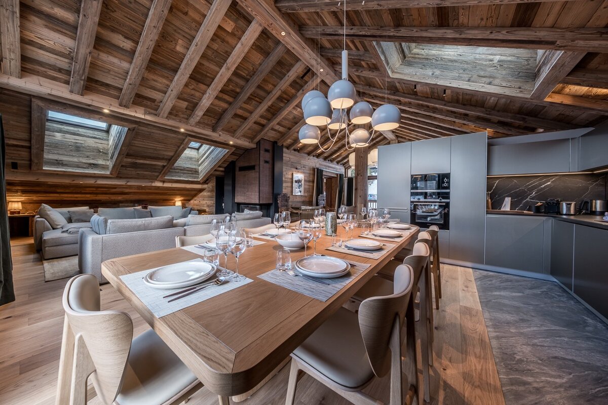 Luxurious chalet interior with exposed wooden ceiling, modern dining area, sleek kitchen, and cozy living space under skylights.