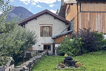 A stone house with wooden shutters stands beside a modern wooden building, with a green yard, stone wall, and mountain in the background.