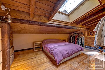 Cozy attic bedroom with warm wooden ceilings, beams, and floor. Features a purple-duvet bed, a bright skylight, and an open clothes rack.