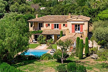 A beautiful Mediterranean villa with terracotta roof tiles, peach walls, red shutters, a swimming pool, and lush green gardens.