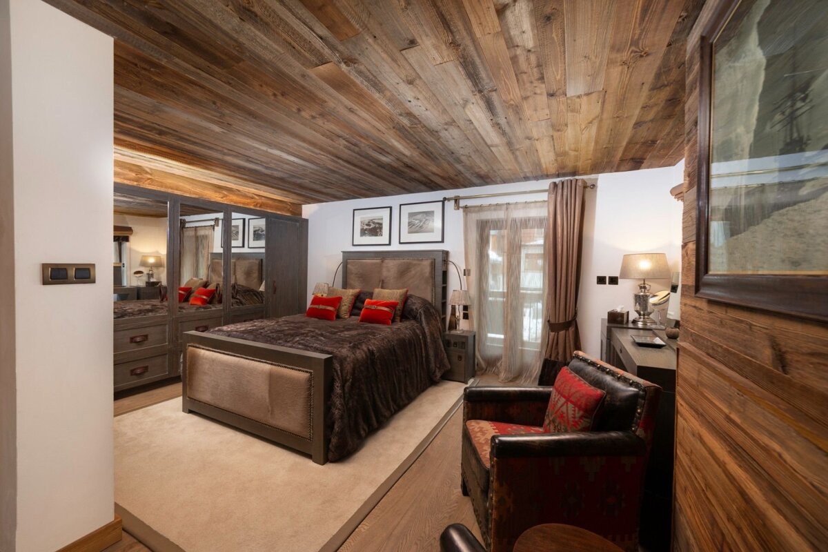 An inviting bedroom features a rustic wood plank ceiling, large bed with dark bedding & red pillows, mirrored wardrobe, and an armchair, creating a cozy atmosphere.