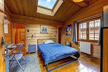 A rustic, wood-paneled bedroom featuring a bed with blue sheets, a skylight, window, desk, and Americana decor like a Route 66 sign.