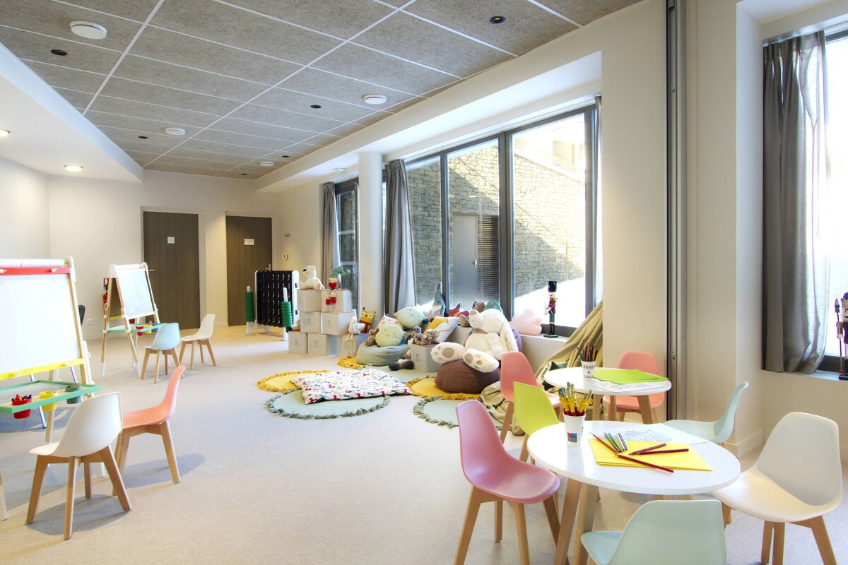 A bright, spacious kids' play room with colorful chairs, small tables, art easels, and a window-side corner piled with plush toys.