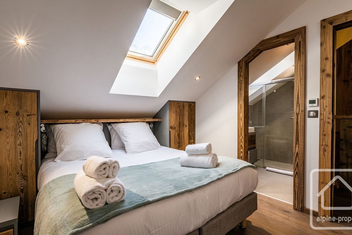 Cozy attic bedroom with a comfortable bed, bright skylight, and warm wooden accents. An ensuite bathroom is visible through a wooden doorway.