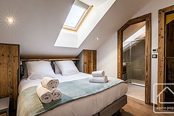 Cozy attic bedroom with a comfortable bed, bright skylight, and warm wooden accents. An ensuite bathroom is visible through a wooden doorway.