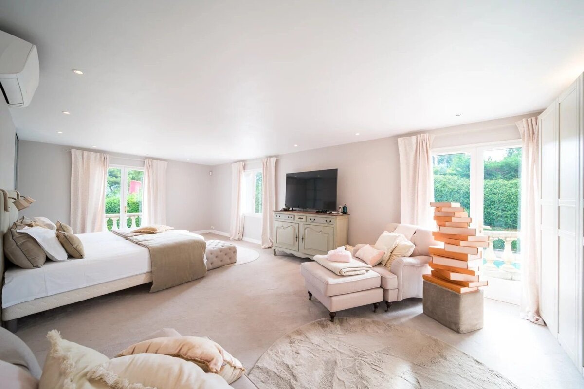 Spacious, elegant bedroom in soft beige tones featuring a large bed, seating area, TV console, and doors opening to a lush garden view.
