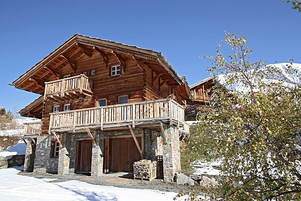 A rustic wooden chalet with stone foundation and balconies sits on a snowy mountain slope under a clear blue sky, with a sparse tree in the foreground.