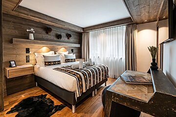 A rustic, wood-paneled bedroom features a large bed with a patterned blanket, a floating nightstand, a desk, and a window with sheer curtains.