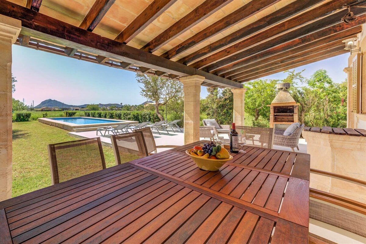 A covered patio with outdoor dining (fruit, wine) overlooking a pool, green lawn, distant mountains, and a BBQ grill.