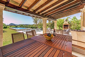 A covered patio with outdoor dining (fruit, wine) overlooking a pool, green lawn, distant mountains, and a BBQ grill.