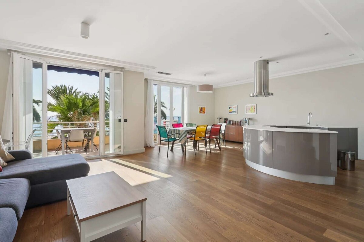 Bright, modern open-plan apartment with a kitchen island, dining area with colorful chairs, and a balcony overlooking palm trees and the sea.