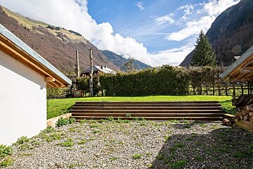 A mountain valley scene features a gravel foreground, wooden steps leading to a green lawn, a hedgerow, and buildings under a blue sky with clouds.
