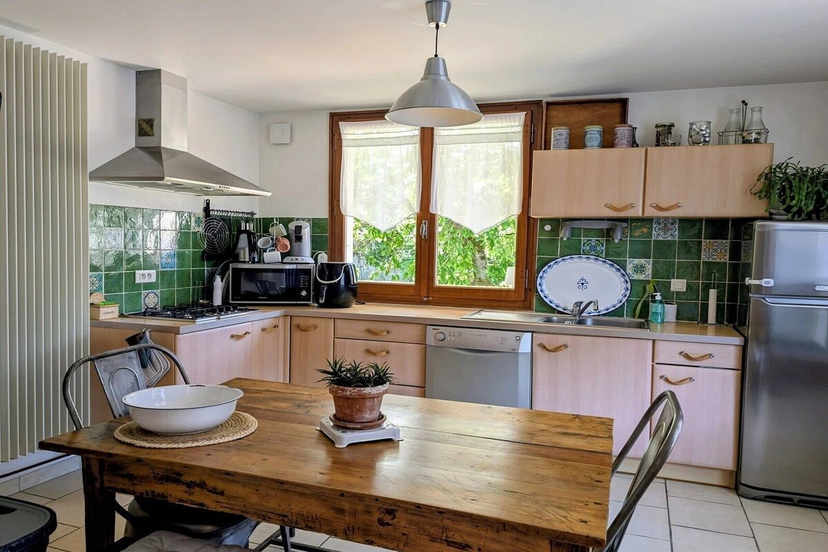 Cozy kitchen featuring green tiled walls, light wood cabinets, and a wooden dining table in the foreground.