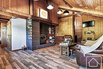 A rustic wooden living room features a dark-tiled fireplace, leather sofa, TV, wood paneled walls/ceiling, and a Stella Artois sign.