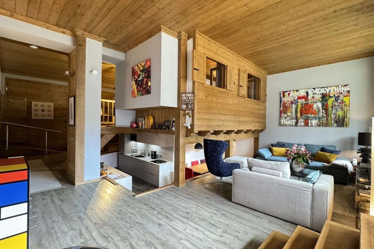 A modern, lodge-style multi-level living room with light wooden elements, a compact kitchen, comfortable seating, and vibrant abstract art.