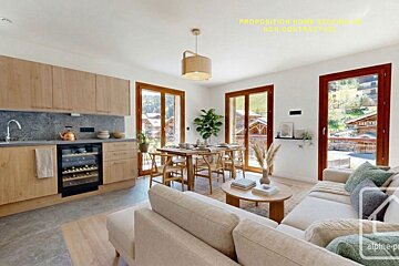 Bright open-plan chalet living space featuring a modern kitchen, dining area, and cozy lounge. Large windows overlook a charming mountain village.