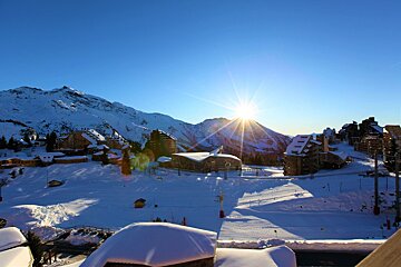 Bright sunburst over a snow-covered mountain village with buildings and ski slopes under a clear blue sky.