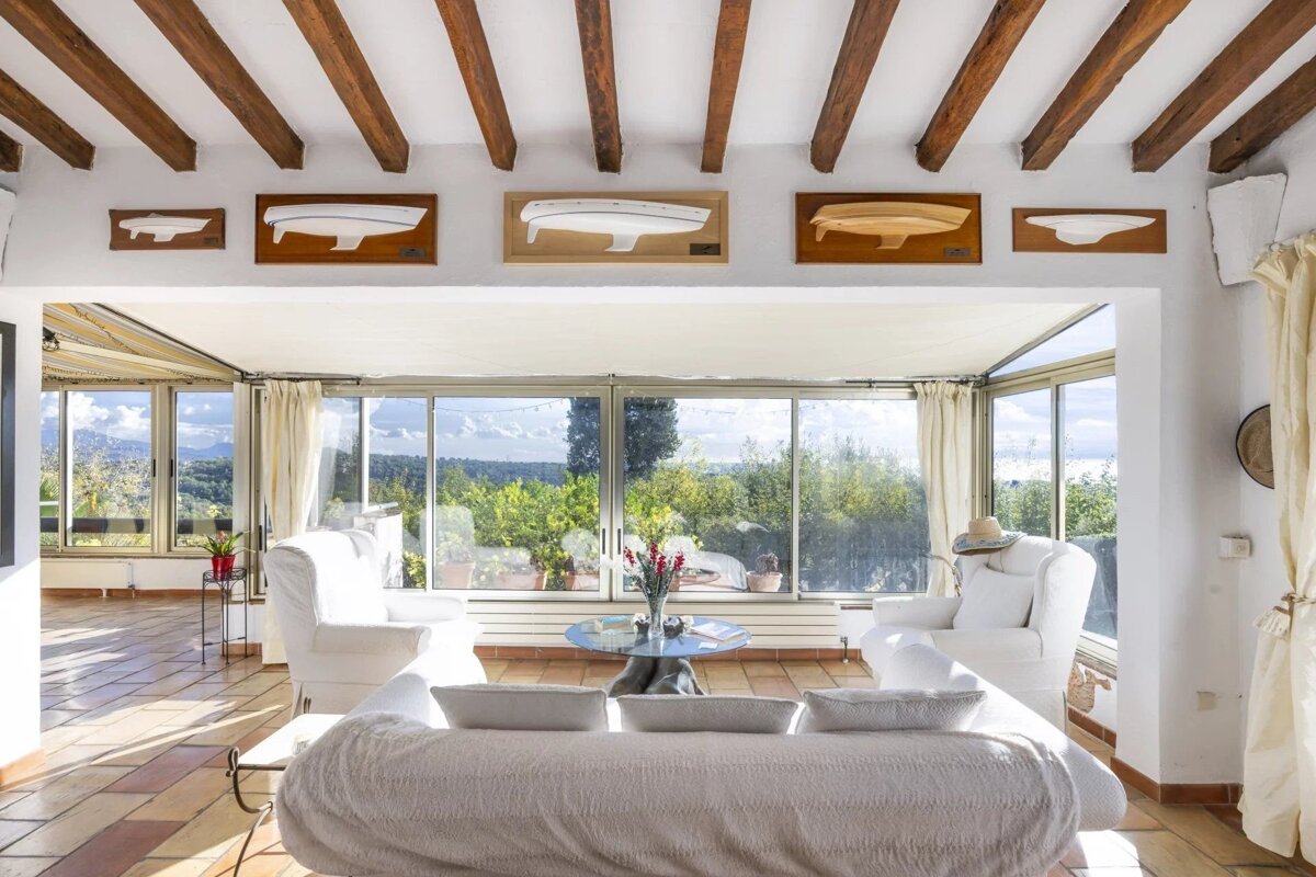 Bright living room with white sofas, exposed wooden ceiling beams, ship hull wall decor, and large glass doors showing a scenic landscape.