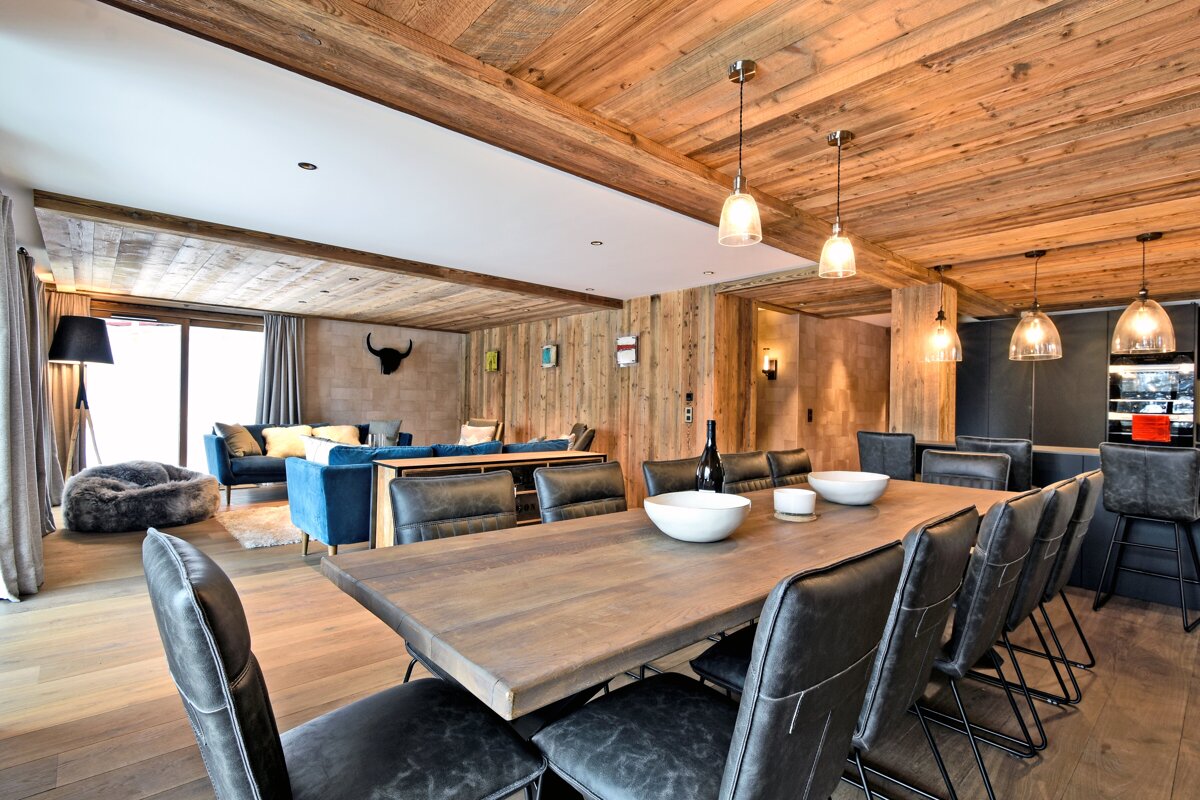 Chic open-plan living and dining area with extensive wood paneling, a long table with leather chairs, and comfortable blue sofas.