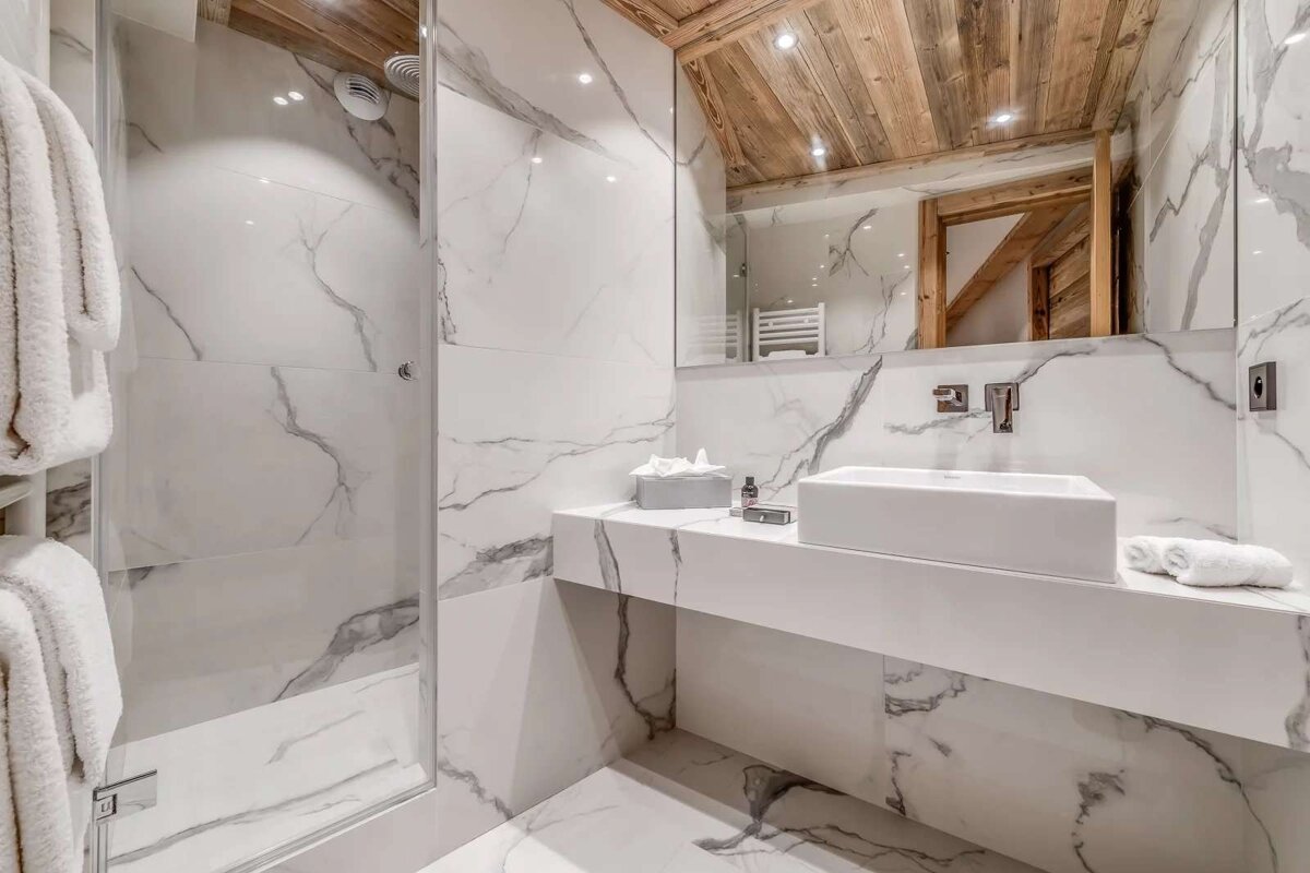 Sleek, modern bathroom with white marble walls/floor, a warm wooden ceiling, glass shower, and a minimalist white vanity with a rectangular sink.