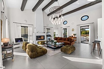 A bright, modern living room with high beamed ceilings, a striking chandelier, fireplace, plush brown/green seating, and patio views.