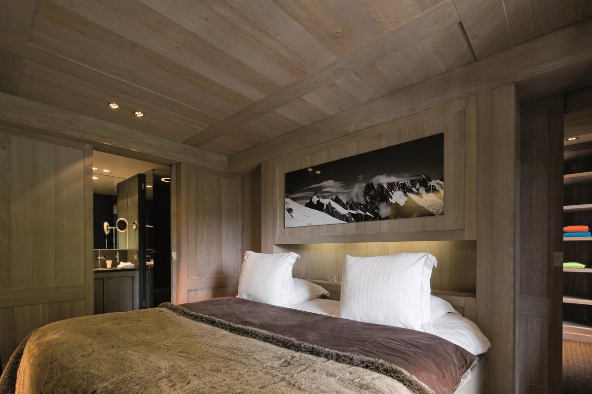 A cozy, wood-paneled bedroom with a large bed, mountain artwork, and an adjoining modern bathroom/wardrobe. Warm lighting creates a inviting atmosphere.