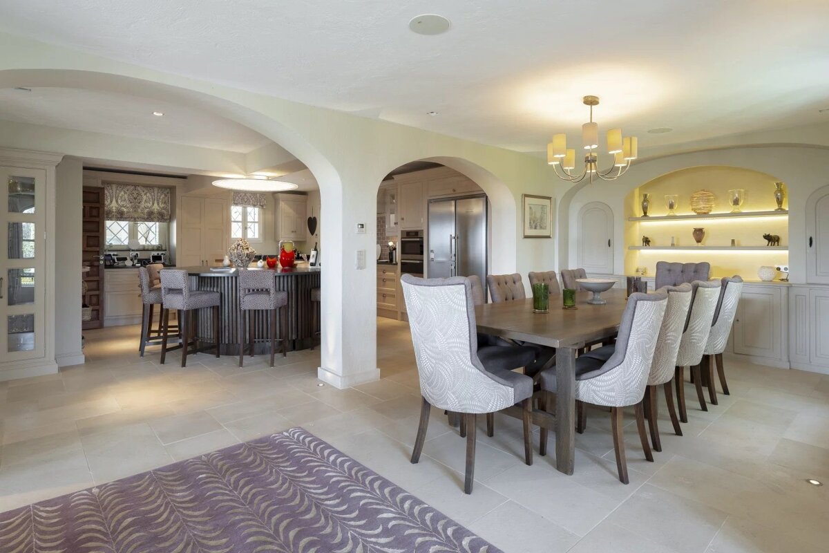 Spacious open-plan kitchen and dining room. Kitchen has an island with bar stools. Dining area features a long table, upholstered chairs, and lit shelves.