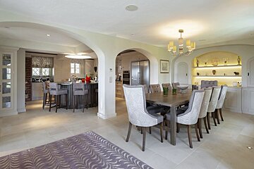 An elegant, open-plan dining area featuring a wooden table with patterned grey chairs, leading through wide archways into a kitchen.