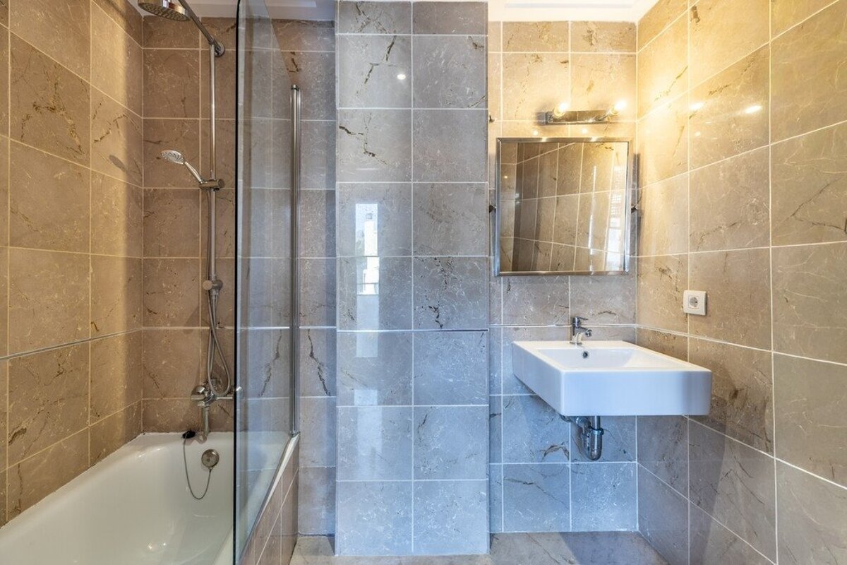 Modern bathroom featuring beige and gray marble-look tiles, a bathtub with a glass screen, and a wall-mounted sink with a mirror.