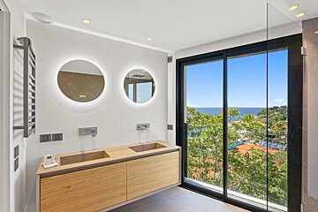 Modern bathroom with a double vanity, round backlit mirrors, and large sliding doors overlooking a scenic ocean and green landscape.
