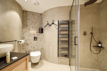 A modern bathroom featuring a curved tiled wall, wall-mounted toilet, heated towel rail, wood vanity, and glass shower enclosure.