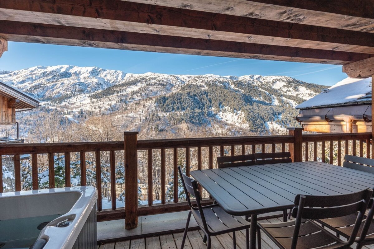 Chalet balcony with a hot tub and dining set, overlooking stunning snowy mountains and a winter forest on a clear day.