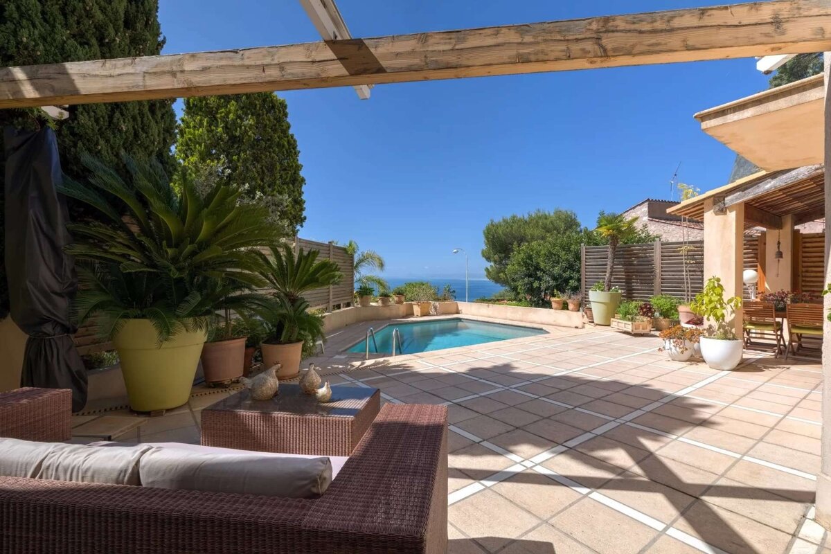 A sun-drenched Mediterranean terrace with a pool, ocean view, and vibrant potted plants. A wooden pergola shades stylish wicker furniture on tiled paving.