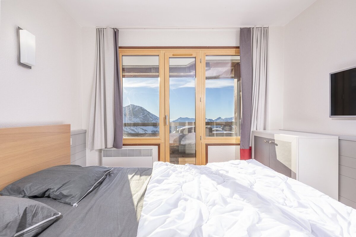 A bright bedroom with a comfy bed, wall-mounted TV, and large glass doors offering a breathtaking view of snowy mountains.
