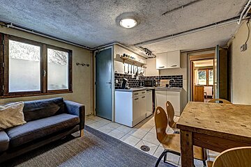 A rustic open-plan room with a kitchenette, blue sofa, and wooden dining table. Windows have frosted lower panes, and ceiling pipes are exposed.