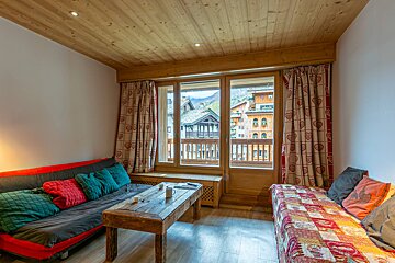 Cozy chalet-style room with a wooden ceiling, large window overlooking a mountain village, two seating areas, and a rustic coffee table.