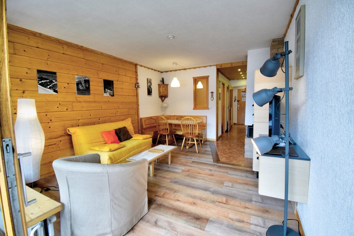Cozy apartment living room featuring a wood-paneled wall, yellow sofa, armchair, and a dining area in the background.