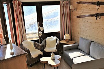 Cozy ski chalet living room with a snowy mountain view, wood-paneled walls, comfortable seating with sheepskin throws, and vintage skis as decor.