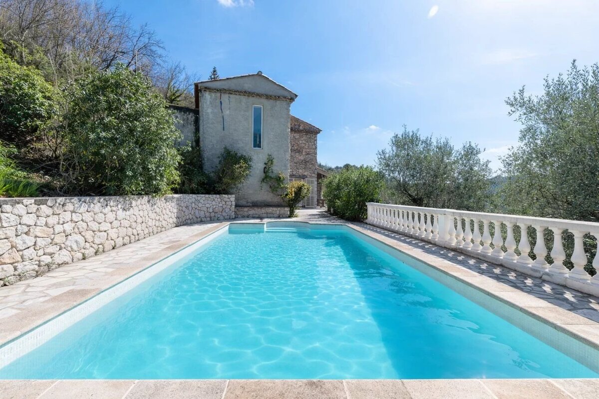 A clear blue swimming pool in a sunny setting with an old stone house, green trees, a stone wall, and a white balustrade.